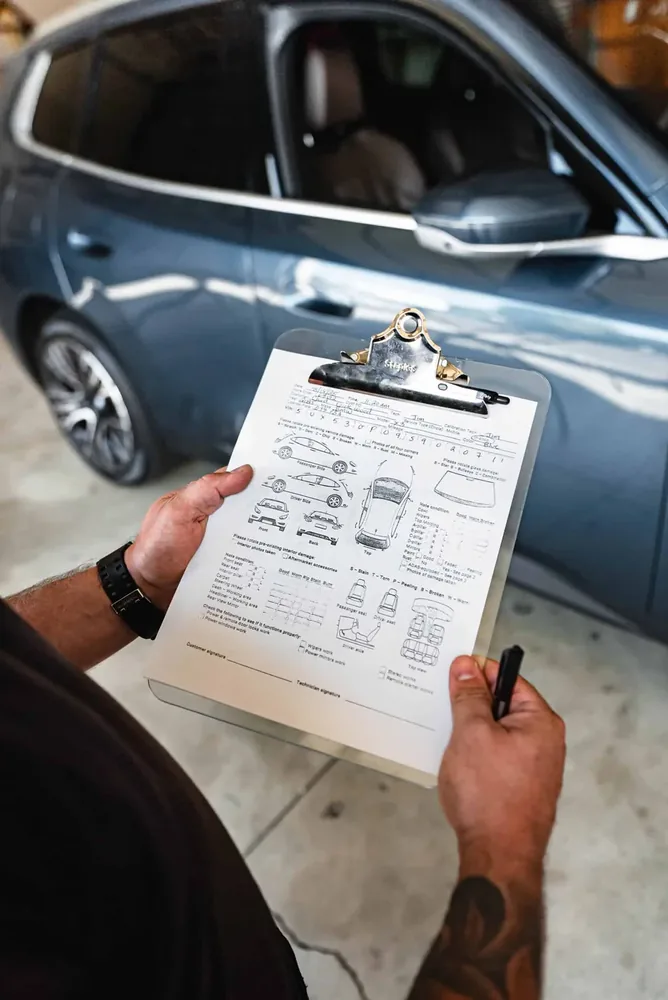 A gloved hand filling in a printed vehicle inspection diagram on a clipboard beside a car
