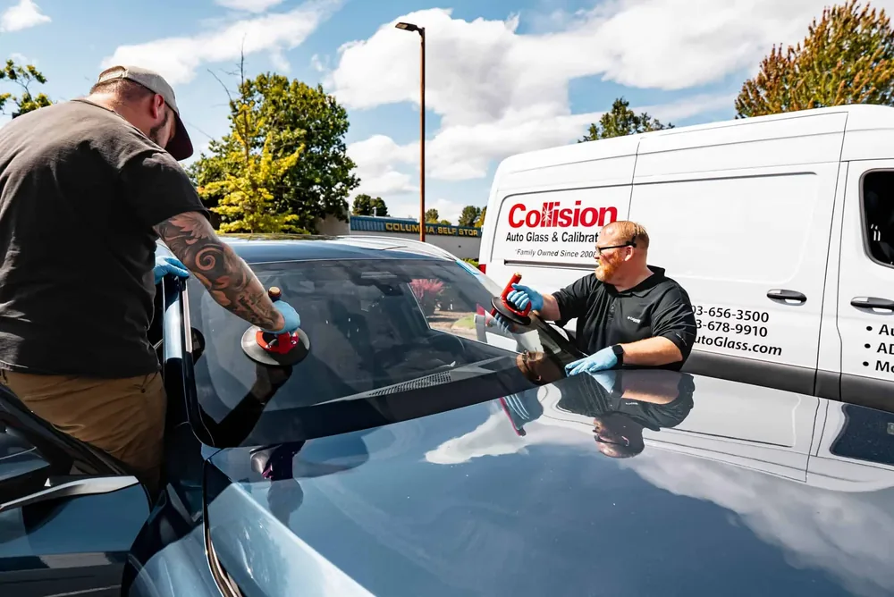 Two technicians positioning a windshield onto a car with the mobile van parked behind